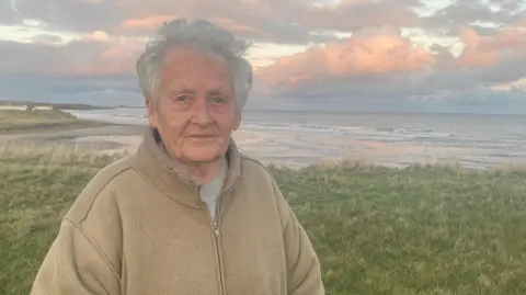 Celia Henry stands with the estuary of the Wansbeck from Cambois beach behind her. She is an elderly woman wearing a brown fleece. The sky is full of pink and grey clouds.