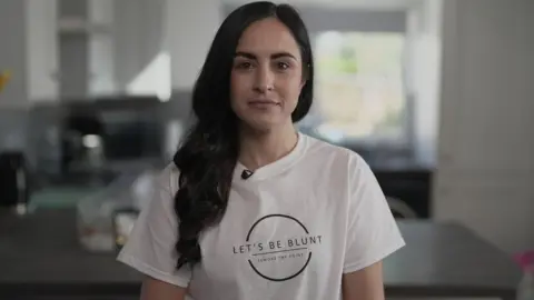 BBC Leanne Lucas, who has long dark hair, smiles at the camera while wearing a white t-shirt with the logo 'Let's Be Blunt - Remove the Point'