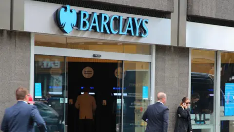 Two men in suits walk outside a Barclays bank on the High Street while one woman is standing outside talking on her mobile phone