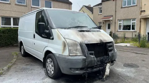 The van has a melted front grille and a cracked windscreen. The registration plate is melted and hanging off.