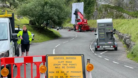 Cliff Road in Cheddar which is closed off with barriers for filming. In the distance there is a lighting rig. A man in a high viz jacket stands to the left of shot looking into the camera.