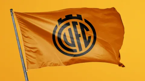 Cambridge United CUFC printed in black on an orange flag with a circle design around it