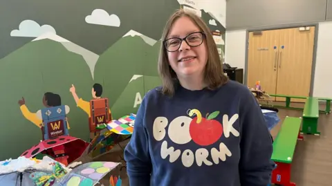 A woman wearing a jumper which says 'book worm'. She is standing in front of a mural which shows two people stood at the foot of a mountain range pointing upwards. there are some green benches behind the woman and a series of colourful, open umbrellas on the floor. 