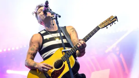 Getty Images Scottish singer-songwriter Callum Beattie is performing on a stage. He is wearing a black and white stripy vest and is playing a yellow guitar.