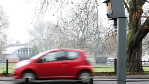PA A red car passes by a speed camera on a road. The car is blurred with the yellow camera sitting on a metal pole behind it. In the background a green space can be seen behind a railing, with a building with a curved roof behind the vehicle. 