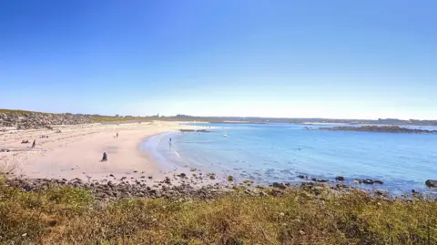 Chris Cumulus/BBC Weather Watchers L’Ancresse - Guernsey. A large sandy beach. There are people walking along it. The the right is blue water. The sky is a bright blue. 