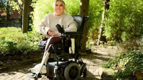 Day One A woman in a wheelchair with one leg and a crutch smiles as she is in a path under some trees in the sun.