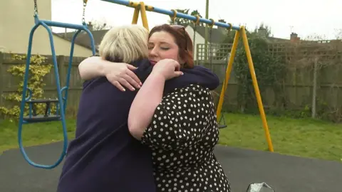 BBC Two women hugging in front of a swing set. There is grass and a wooden fence surrounding the swing set and there are houses in the background above the fence.