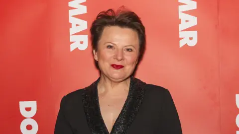 Getty Images Monica Dolan wearing a black top and smiles at the camera. She is standing in front of a red background. 