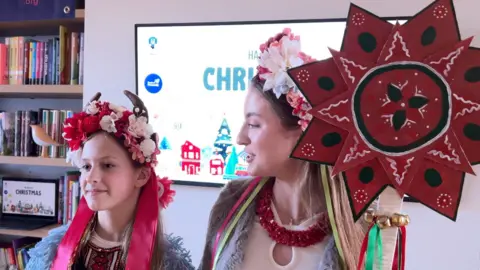 Two young women stand on stage wearing garlands of flowers in their hair. The younger one on the left also has a headband decorated with goats horns to symbolise the animal she is playing.
