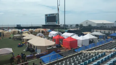 MapAction A temporary emergency response base set up inside a stadium near the coast, with multiple tents, equipment, responders and rows of empty seats visible.