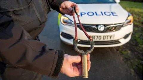 A police officer stands in front of a patrol car and holds a catapult.