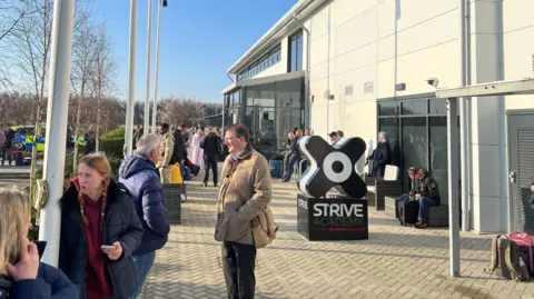 Groups of people stand outside Strive Academy in the sunshine. There is a police car in the background. 