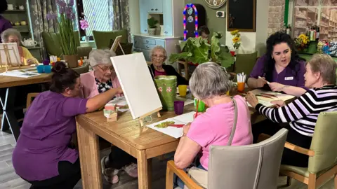 Cathy Killick/BBC A group of four elderly women sit around a table painting on canvases. Two women in purple carer uniforms crouch next to the table as they speak together.