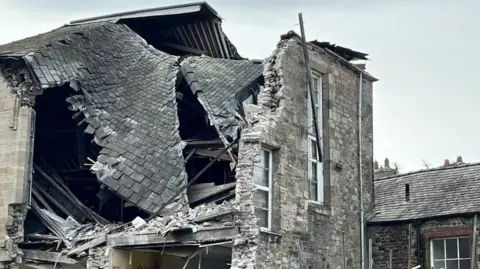 BBC/Andy Greenwood A grey-stoned three-storey building that has collapsed. A wall is missing with brickwork and metal poles hanging precariously.