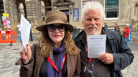 BBC A woman in large sunglasses and a large-rimmed hat as standing beside a man with grey hair and beard in the street as they hold fliers for their play