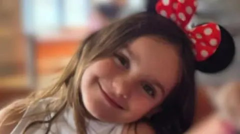 A girl tilts her head to the side as she smiles at the camera. She has long brown hair and is wearing Minnie Mouse ears on her head.