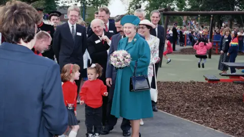 Getty Images Queen Elizabeth in the same blue/green outfit smiling at crowds. She's standing in Pallister Park. Children can be seen on the swings in the background.