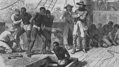 Black-and-white historical engraving showing several captive people on the deck of a ship. Some have leg shackles and are being restrained by crew members, while others sit or kneel nearby. One captive person is shown emerging from a hatch in the deck. The scene depicts the confinement and control of enslaved people aboard a slave ship during the transatlantic slave trade.