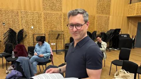Katy Lambert/BBC A middle-aged man with grey hair and a black polo shirt smiles into the camera. He has sheet music in his hands. Two people are sat on chairs behind him in what appears to be a rehearsal area.