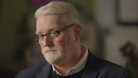 Jim Pearson, a man with grey hair and a grey beard, looking at the camera. He is wearing glasses, a white shirt, a black jumper and a dark blue jacket.