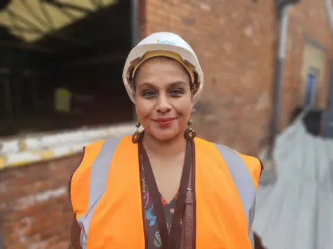 A woman wearing a white hard hat and orange high-vis vest. She has large earrings and a brown bag strap over her shoulder.