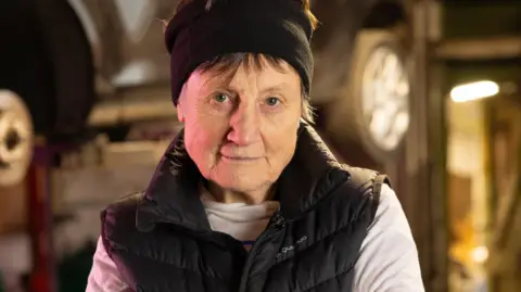 Historic England A person standing with arms crossed in an automotive repair shop, wearing a black headband, black quilted vest, and white long-sleeve shirt. Behind them, a car is elevated on a hydraulic lift, surrounded by workshop tools and equipment under warm lighting.
