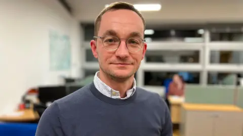 A smiling Tom Abell looks directly at the camera. He is wearing glasses, a blue jumper with a striped shirt underneath. He is standing in an office, with desks and computers behind him. 