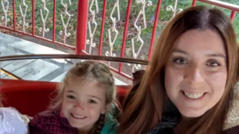 A woman with long dark hair sits on the right of the image smiling into the camera. To her right is a small girl with blonde hair.