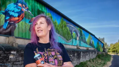 A woman stands in front of a colourful mural and holds a spray can. She has purple hair and glances off to the side