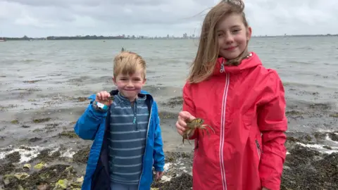 Image shows two children smiling at the camera holding crabs. The girl on the right is wearing a pink waterproof jacket. Her hair is blowing in the wind and she is smiling at the camera. She is holding a big crab. Her brother on the left is wearing a blue waterproof jacket and a blue striped quaterzip. He has blond hair, is smiling and holding a small crab. Behind them, you can see water and Portsmouth. 