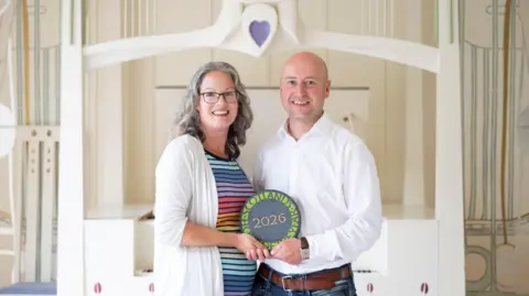 Emily and Robert Haistans stand side by side in the House for an Art Lover, holding their award, a round plaque that reads “2026,” while facing the camera.
They are positioned in a light, decorative interior with pale wood panelling, curved architectural details, and a heart motif above them.