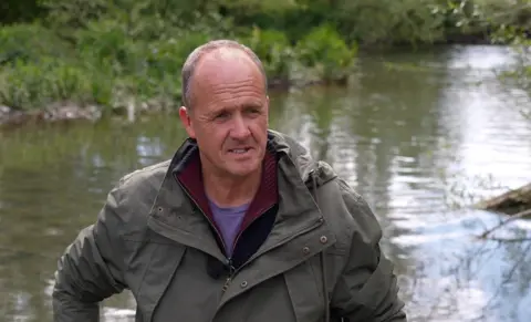 A middle-aged bald man wearing a waterproof jacket. He is standing next to a river.