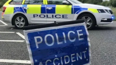 A blue police sign which says 'Police, Accident' propped up in the middle of the road. Behind it is a parked police car.