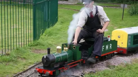 Fancott Miniature Railway A miniature steam locomotive travelling on a narrow-gauge railway through a grassy outdoor area. A person is seated on and operating the locomotive, and white steam is rising from the engine's chimney and lower pipework.
