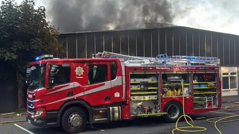 Shropshire Fire and Rescue Service A fire engine is outside a factory warehouse building with large clouds of thick, black smoke above it. There is a yellow hose leading from the engine.