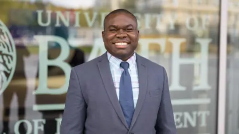 University of Bath Dr Teslim Bukoye smiling at the camera wearing a dark grey suit in front of a window with University of Bath written on it