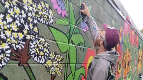 A street artist wearing a purple baseball cap and grey hoodie top spray paints a flower onto a brick wall.