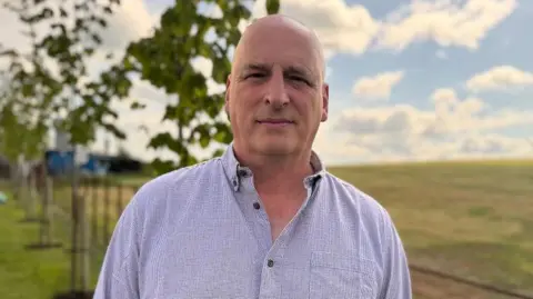 John McClenaghan pictured on a farm. He is bald and wearing a purple lined shirt with brown buttons. He is looking directly into the camera. 