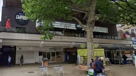 The exterior of HMV Empire in Coventry, a venue above a row of shops which has several large HMV signs. People walk past on the street.