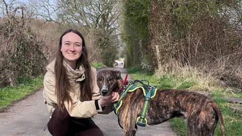 Animal care assistant Amy Robinson, who has long brown hair, with Charlie, a brindle and white lurcher dog wearing a green and yellow harness. Amy is crouched down on a country road cuddling Charlie. Amy is smiling. Charlie is facing the camera.