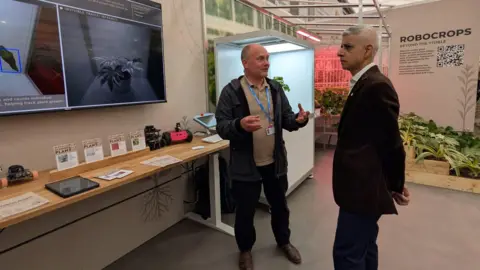 Two men standing next to a black screen which shows a black and white photo of a plant in a pot. A table below it shows three labels saying "plant" with other wording below. Tablets are also on the table. Next to it is a lit-up case with a plant in it and on the far wall it says "Robocrops" with more text and a QR code underneath it. One of the men has a lanyard on and his hands outstretched while the other man, wearing a suit, is looking inquisitively at the screen.