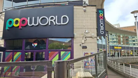 The entrance to popworld, which is a colourful facade with some seating outside. To the right in the background is the entrance to Walkabout.