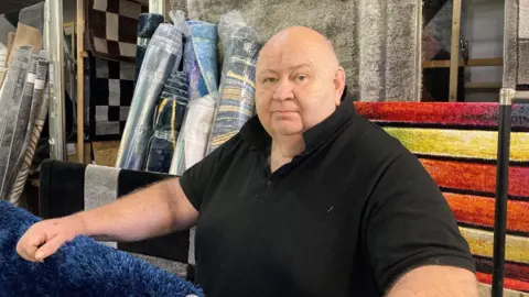 A man in a black polo shirt leans on a rug display, in front of more rugs at his market stall.