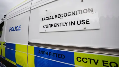 ANDREW MATTHEWS/PA The image shows the side of a van with a white sign on it saying "Facial recognition currently in use". The word "Police" can be seen written further along near the front in blue.