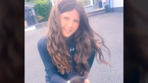 Family handout A young girl with long brown hair smiles into the camera. She is kneeling down and holding a brown kitten. She is wearing a black tracksuit top and joggers.