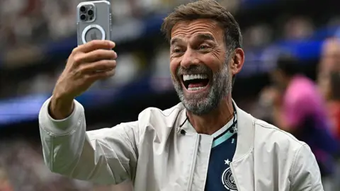Jurgen Klopp smiling while holding a mobile phone in front of his face