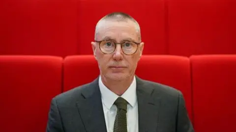 PA Media NUH chief executive Anthony May is pictured looking at the camera. He is stood in front a group of red seats and wearing a white shirt, dark grey blazer and olive tie