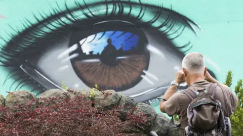 Bristol Business Improvement District A close up of a large brown eye painted on a wall with a turquoise background. The eye is reflecting a person in the iris. A man is standing taking a photo of the eye with his back to the camera and against a wall. He has grey hair and a rucksack