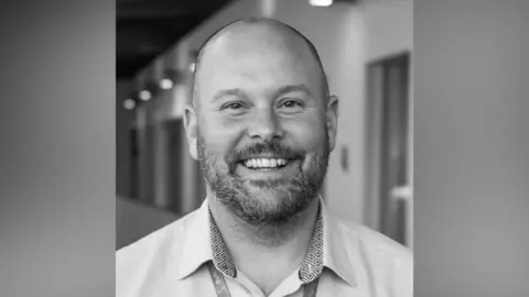 Contributed A black and white image shows Lee Mandley, with shaved head and a beard, smiling at the camera. He is wearing a light-coloured open-neck shirt.